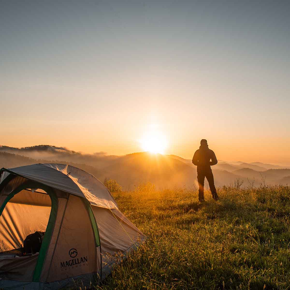Silhouette of a person by a tent at sunrise, enjoying a peaceful camping vacation in nature.