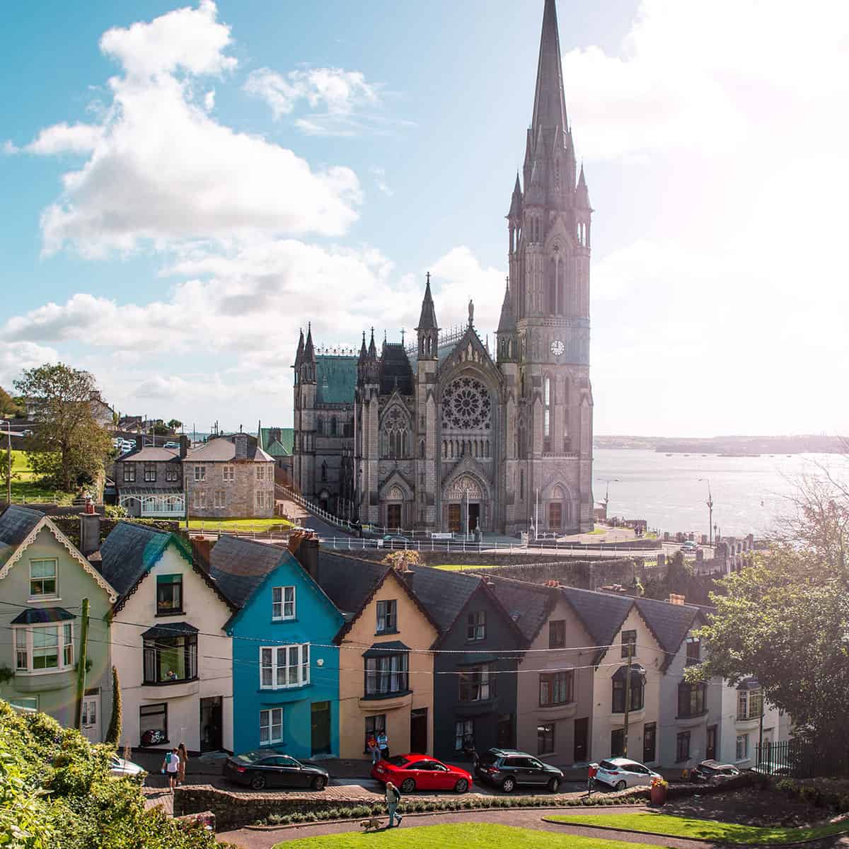 St. Colman's Cathedral towering over colorful houses in Cobh, Ireland, against a bright sky and waterfront.