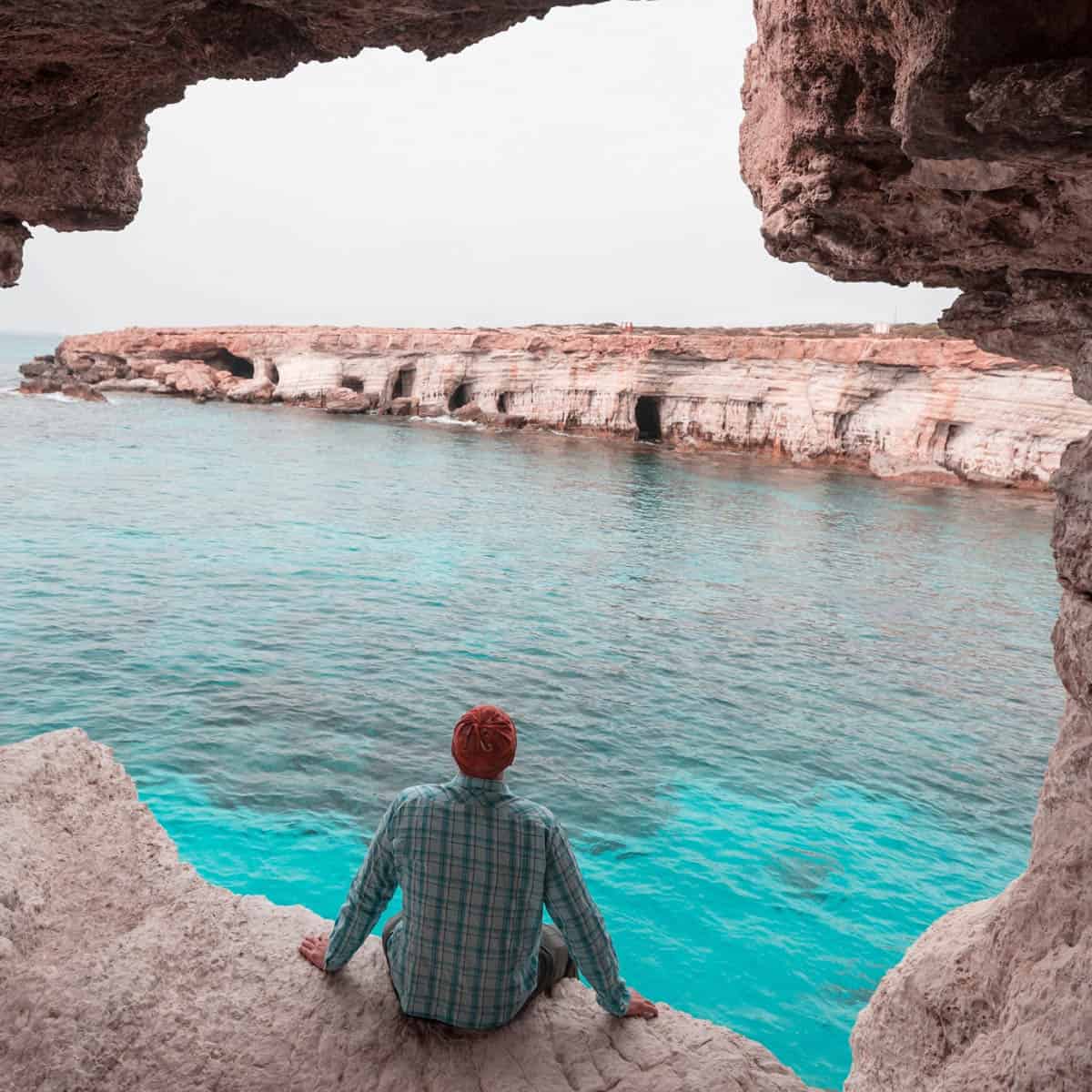 Person enjoying a tranquil view of the turquoise sea and rocky coastline in Cyprus, framed by a natural cave.