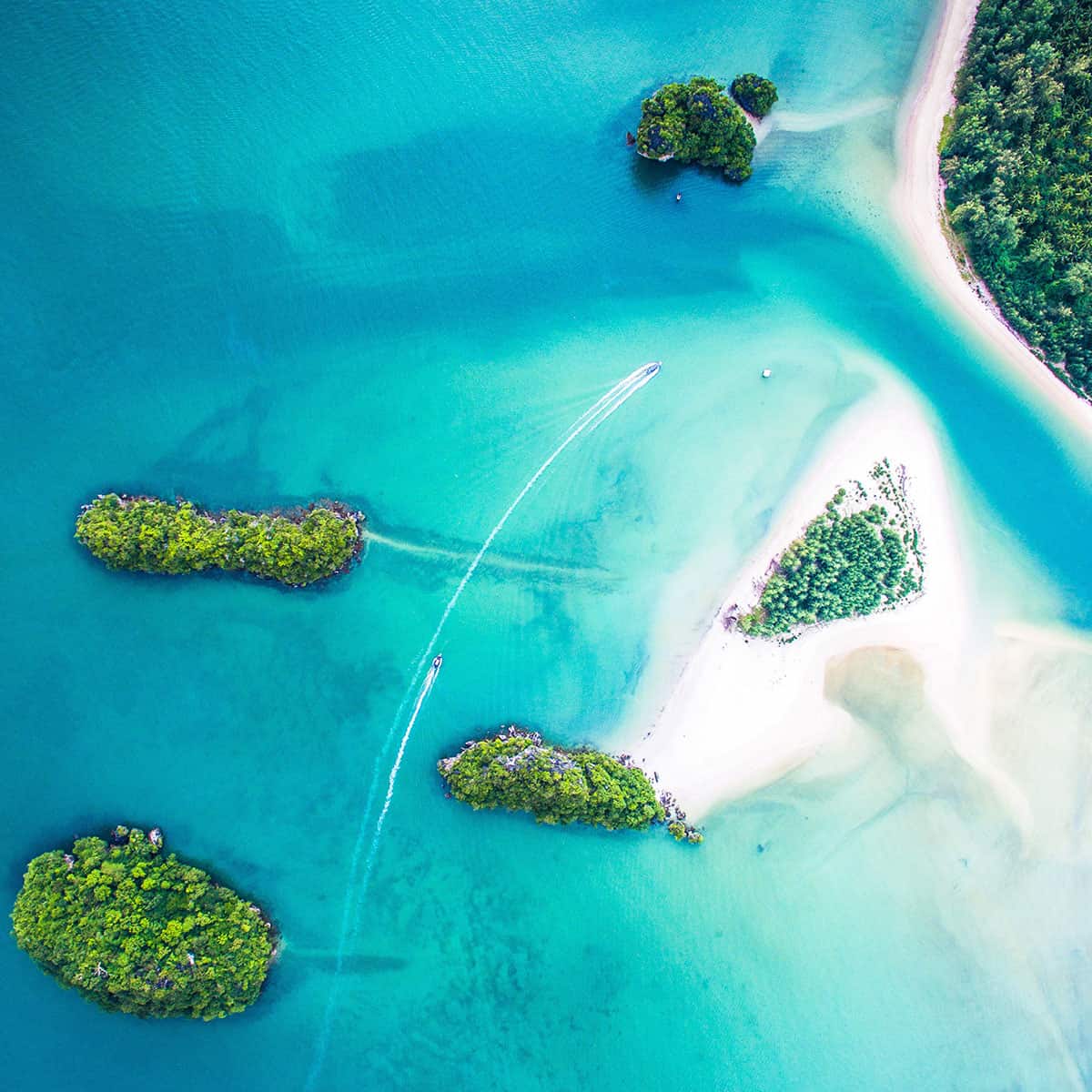 Aerial view of a tropical beach with turquoise waters, small islands, and a boat leaving a wake in the serene landscape.