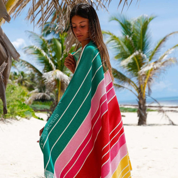 Model wearing colorful Ocean Nile Moss beach towel on a sandy beach surrounded by palm trees.