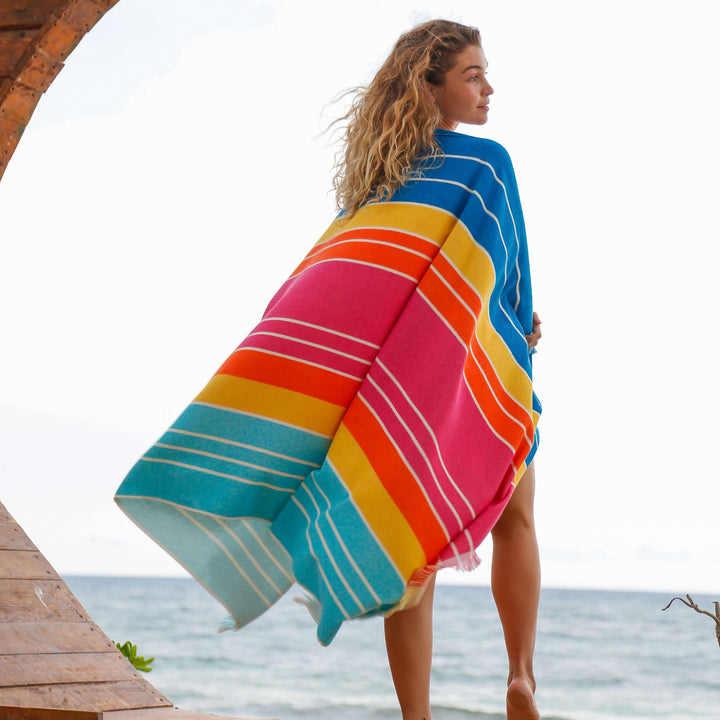 Woman draped in an Ocean Lagoon Aqua Beach Towel, vibrant striped design with a beach backdrop.