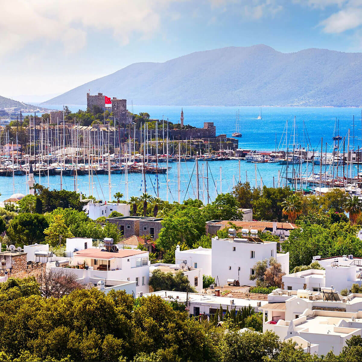Stunning view of Bodrum's harbor with sailboats, lush greenery, and mountains in the background.