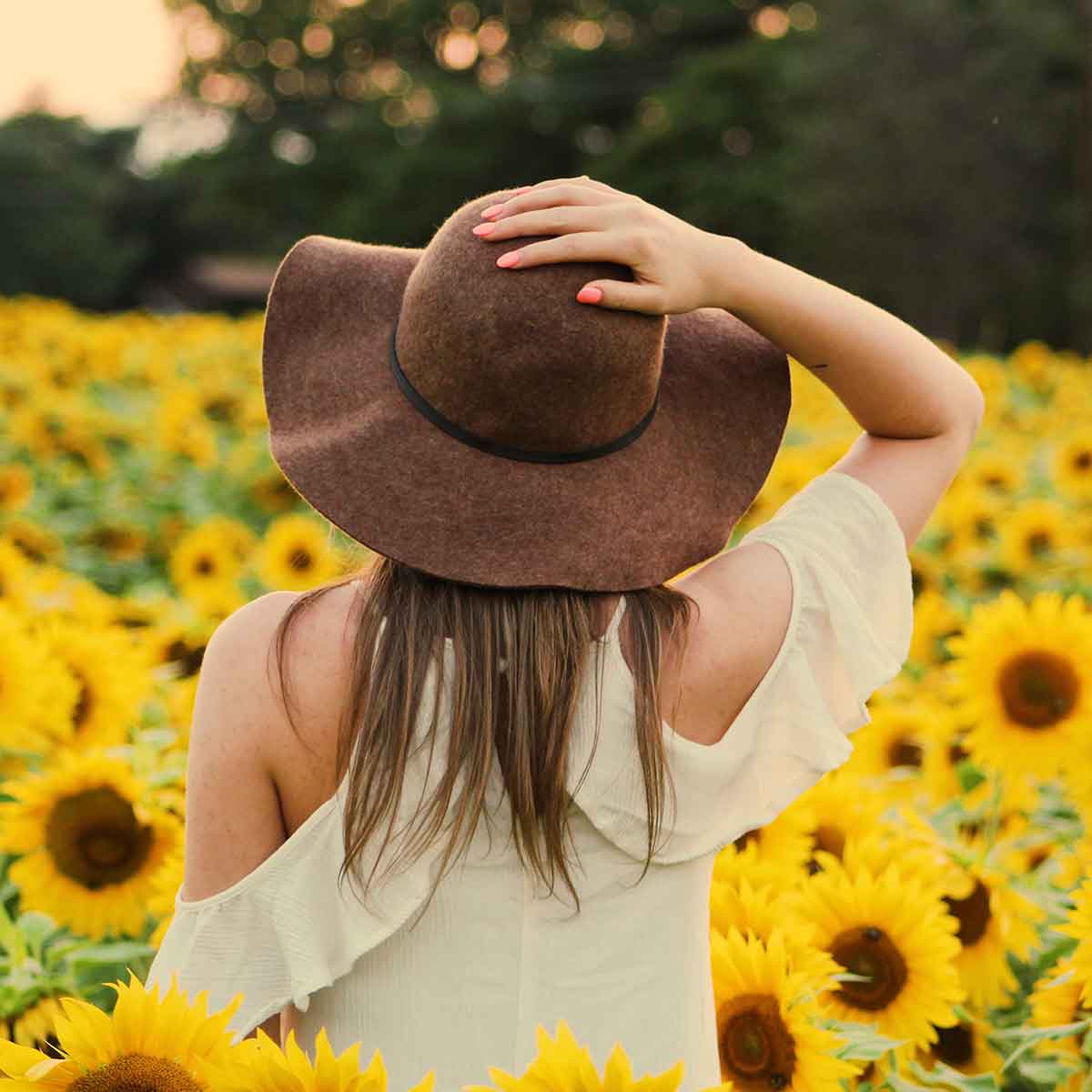 Woman wearing a brown hat, standing in a field of sunflowers, enjoying a sunny day outdoors.