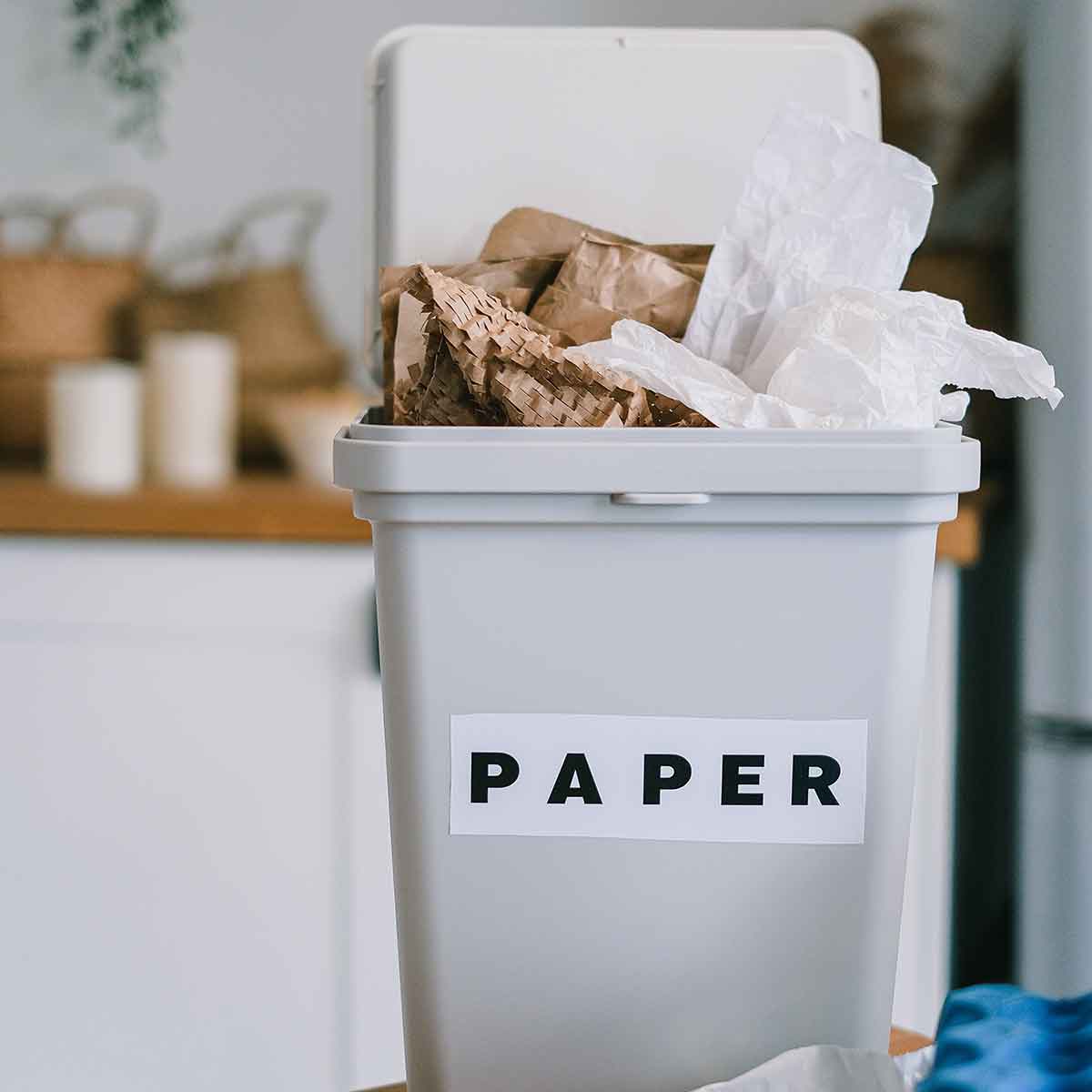 Gray recycling bin labeled 'PAPER' filled with crumpled paper and cardboard.