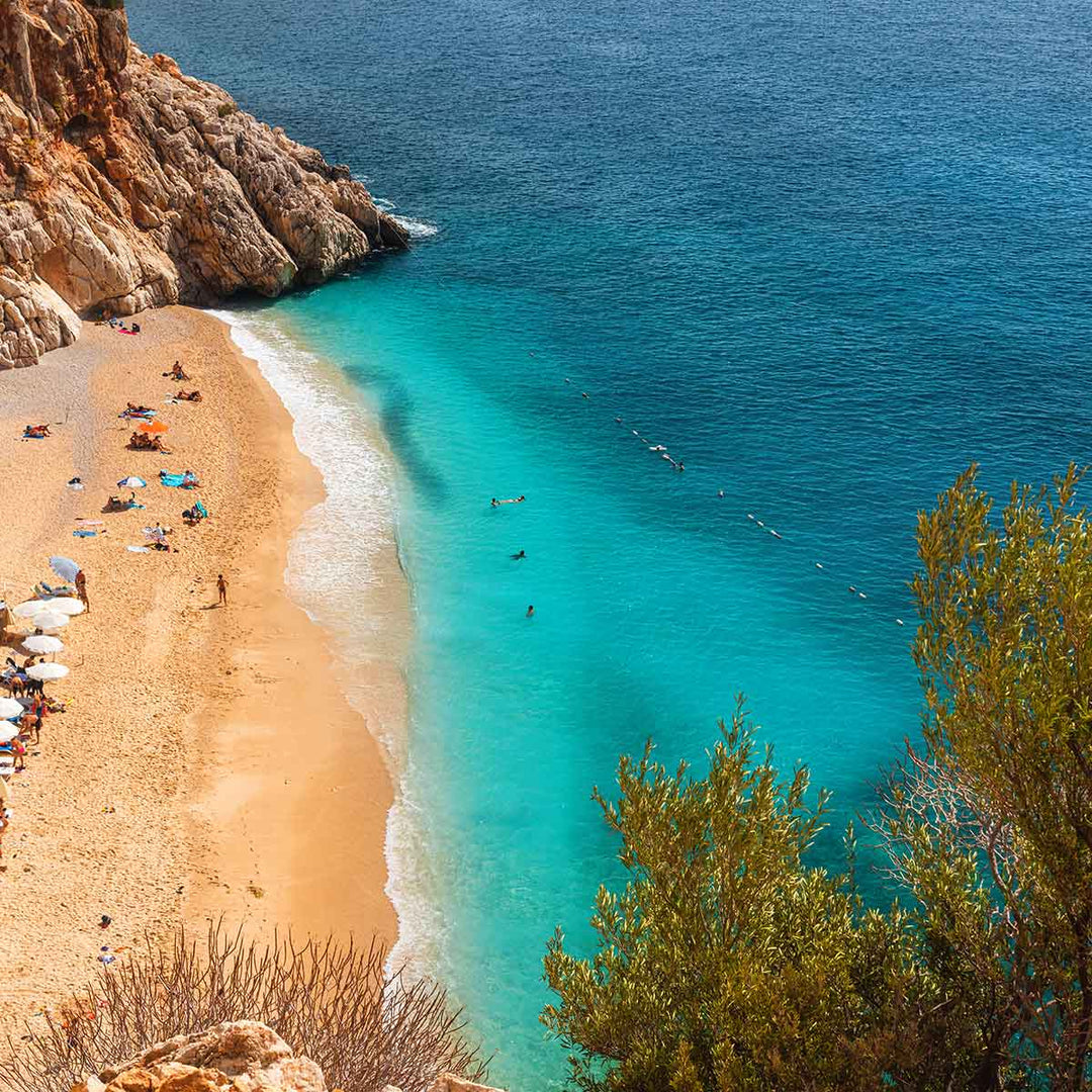 Aerial view of a sunny beach in Kaş, Turkey, featuring turquoise waters and people enjoying the sand.