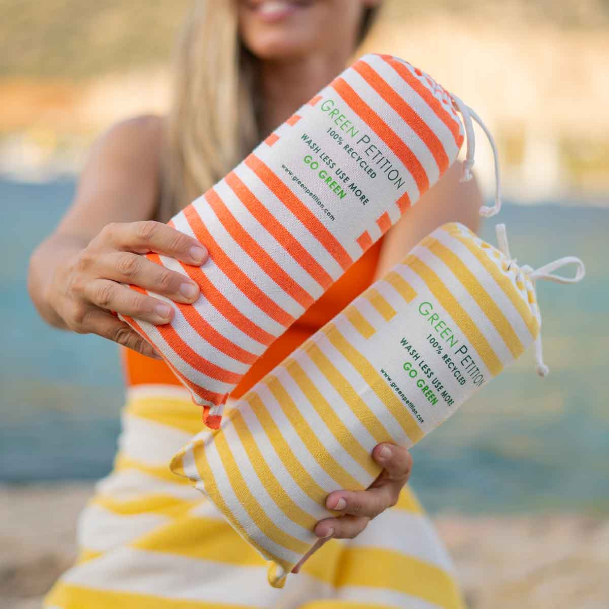 Woman holding two striped sustainable towels from Green Petition by the beach, promoting eco-friendly products.