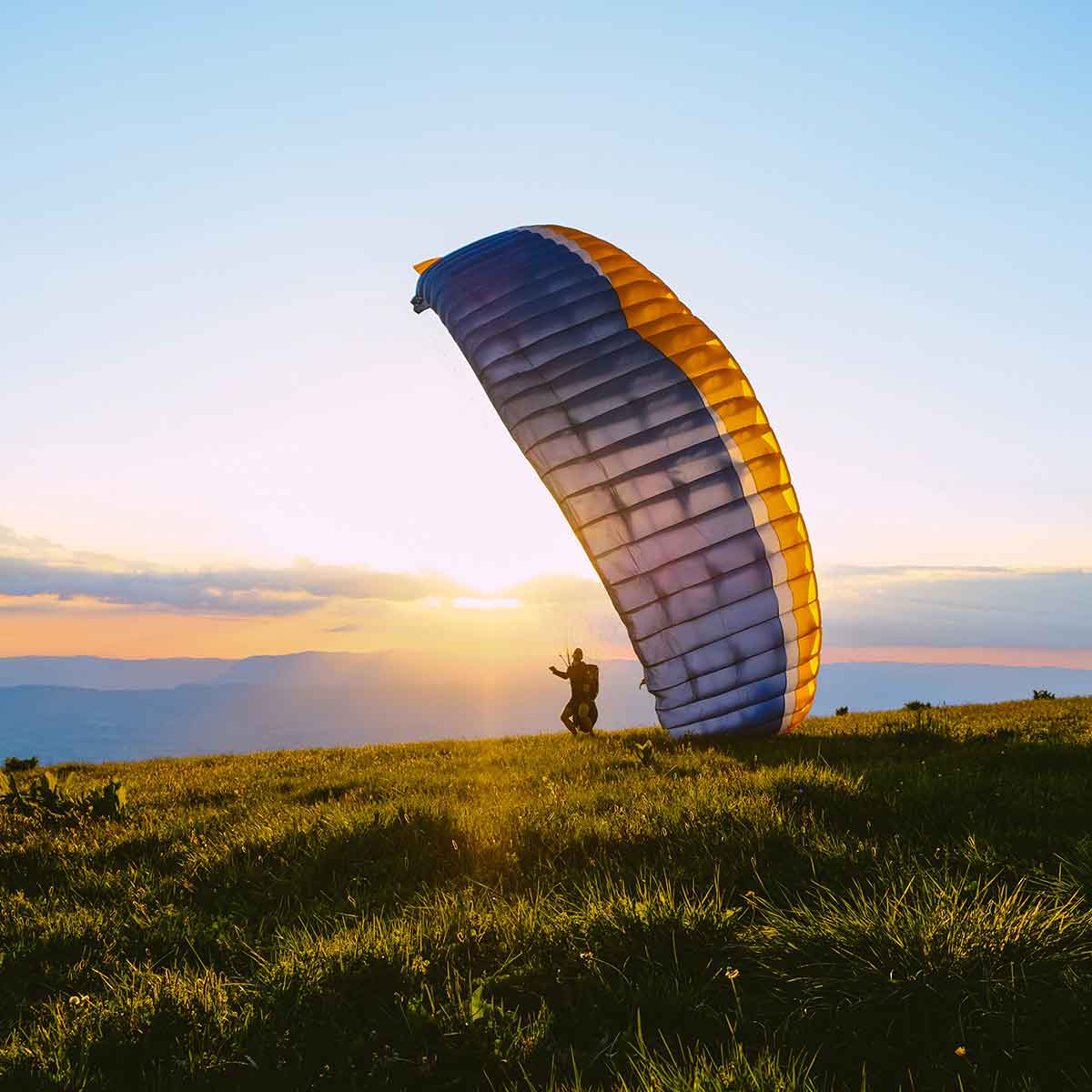A paragliding parachute glides in the sunset, with a silhouette of the pilot preparing for flight.