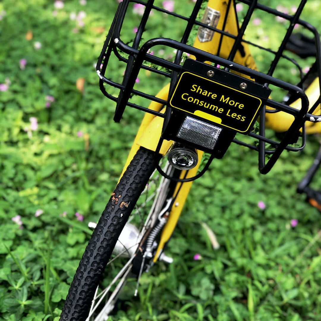 Yellow bicycle with a sign that reads 'Share More Consume Less' on its front basket, surrounded by green grass and flowers.