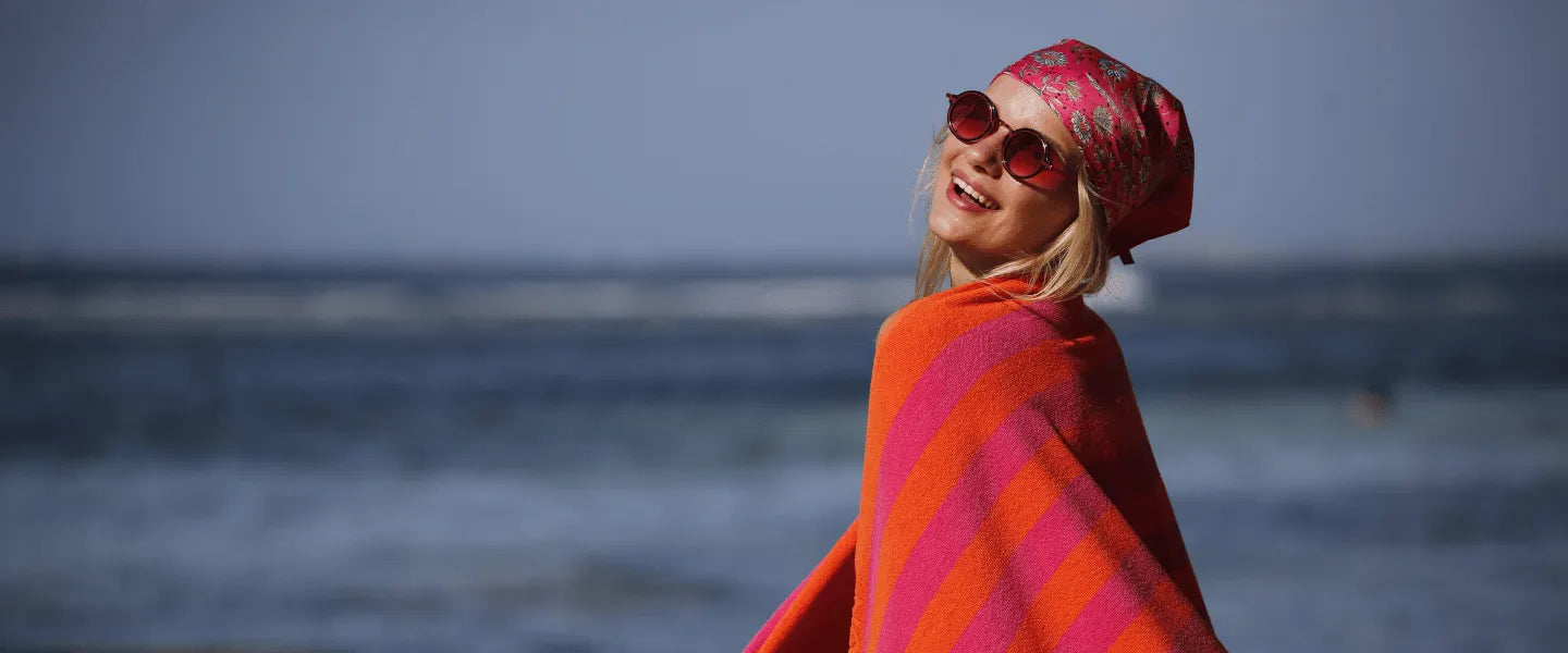 Woman wrapped in a red and fuchsia color striped Green Petition Maris Ruby beach towel on a beach