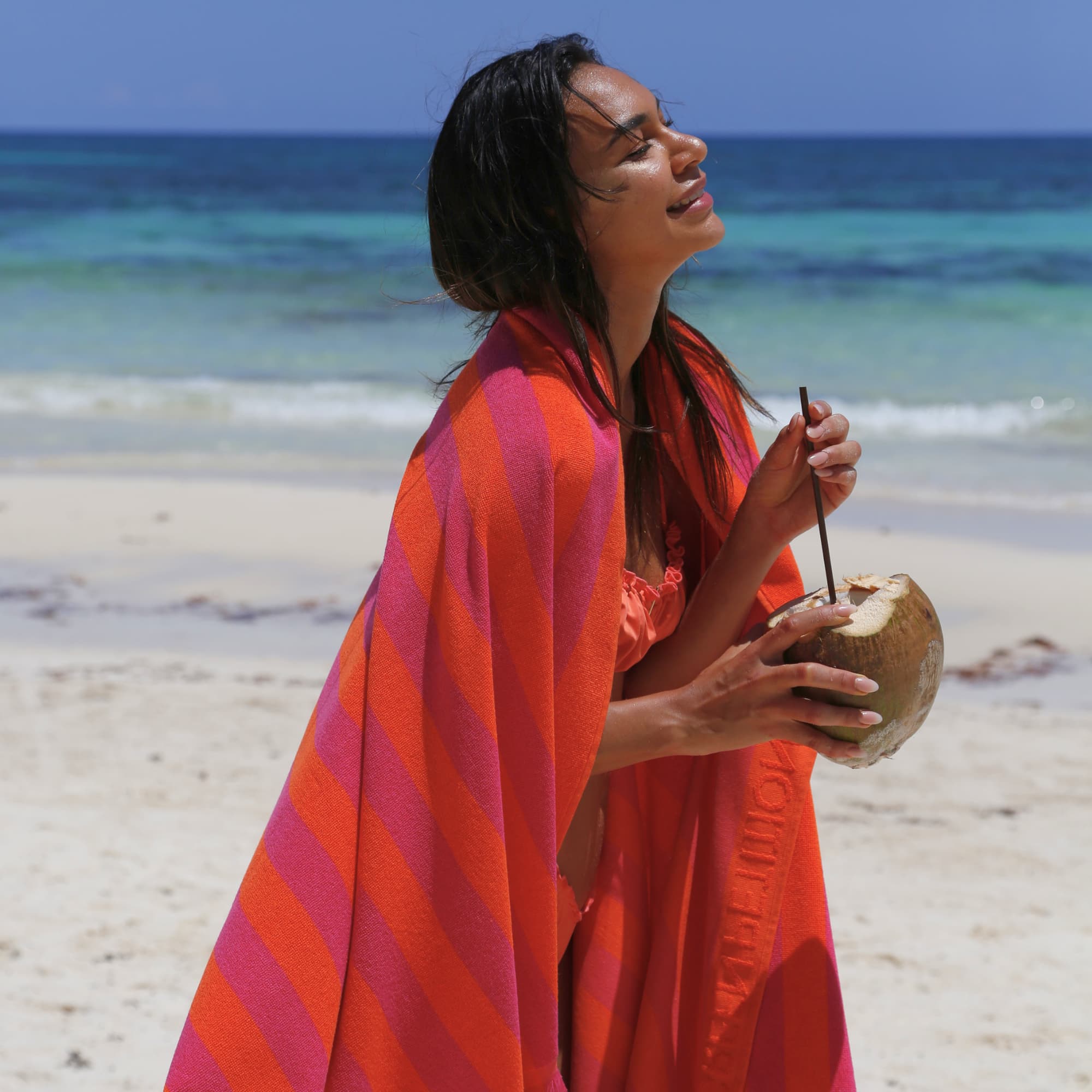 Woman wrapped in a red and fuchsia color striped Green Petition Maris Ruby beach towel on a beach