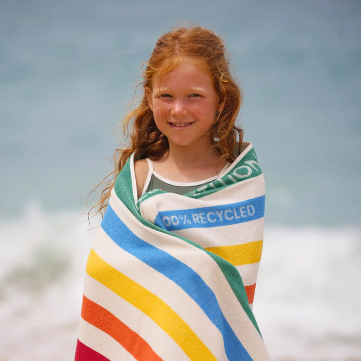 Child wrapped in a colorful striped towel with '100% Recycled' text on a beach.