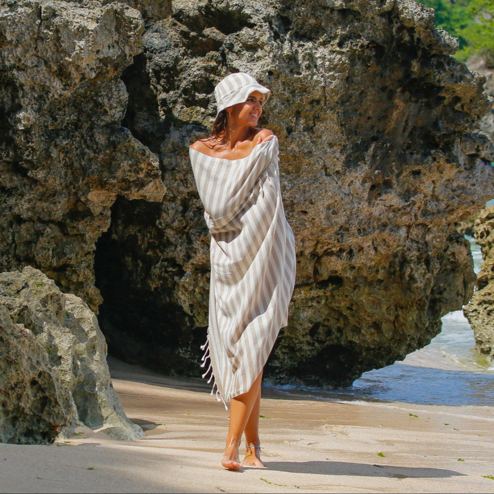 A woman wrapped in a "Green Petition Mare Sand Beige" towel, standing on the beach with rocky cliffs and ocean in the background