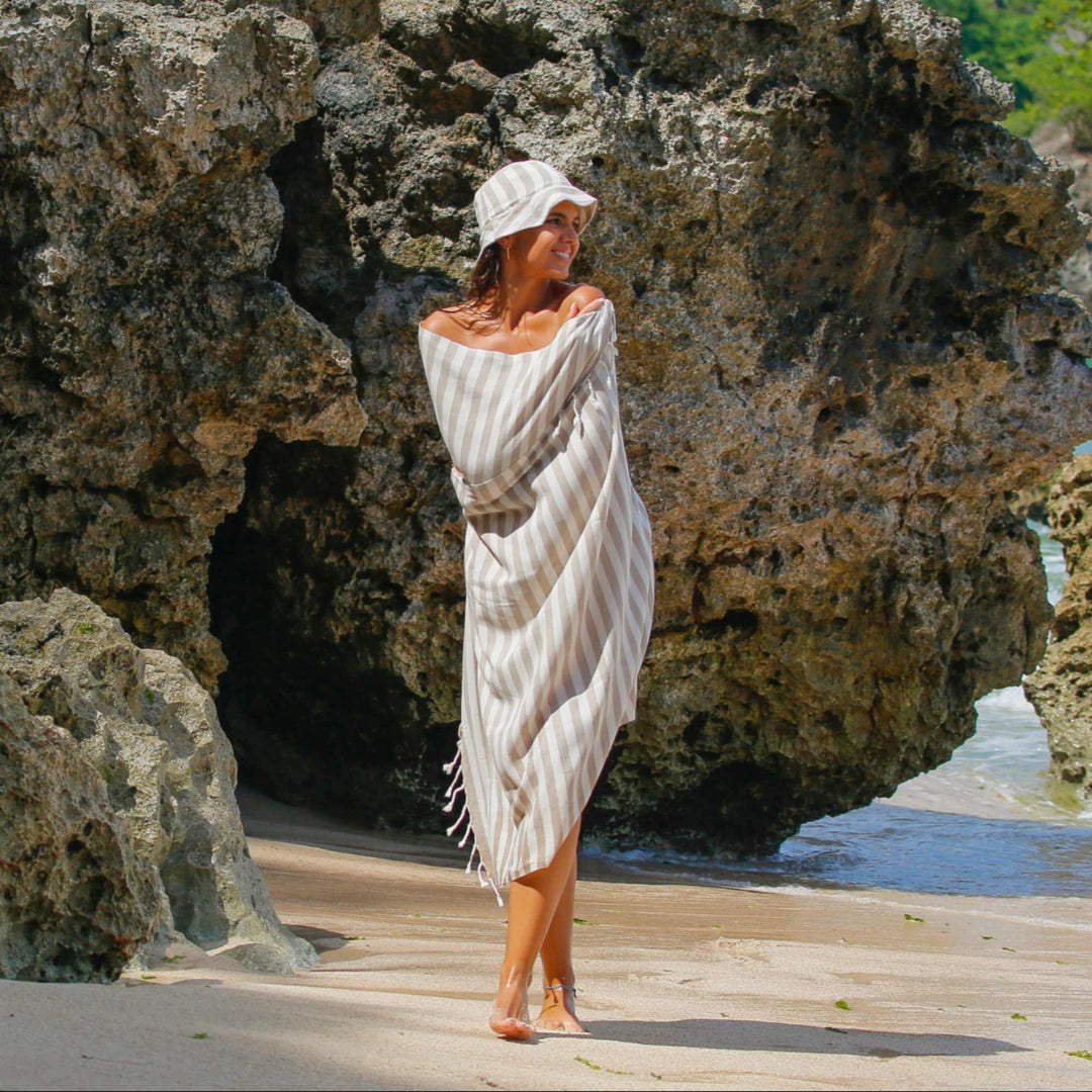 A woman wrapped in a "Green Petition Mare Sand Beige" towel, standing on the beach with rocky cliffs and ocean in the background
