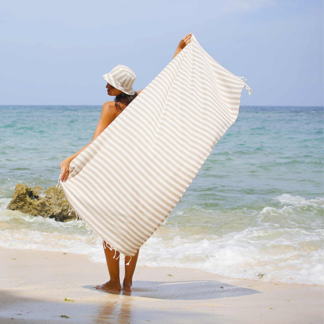 Person holding a white textured "Green Petition Mare Sand Beige" towel on a beach with ocean in the background