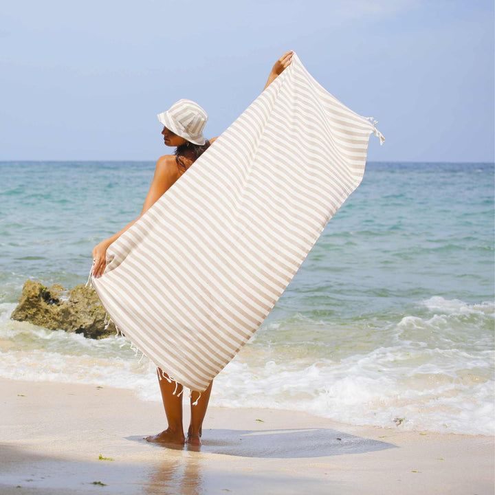 Person holding a white textured "Green Petition Mare Sand Beige" towel on a beach with ocean in the background