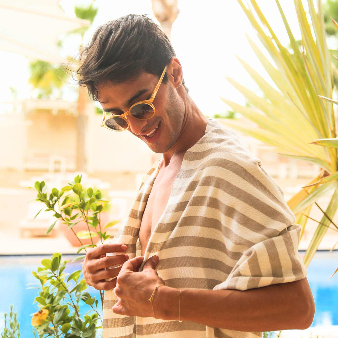 A man wearing sunglasses and a striped "Green Petition Mare Sand Beige" towel by plants and a pool