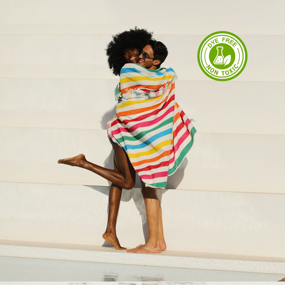 A colcored beautful woman who has fluffy curly hair and hugg the her boy frend. Both of them has wrapped in a colorful striped Green Petition 100% Recycled towel with a 'Dye Free' and 'Non Toxic' logo on a light background.