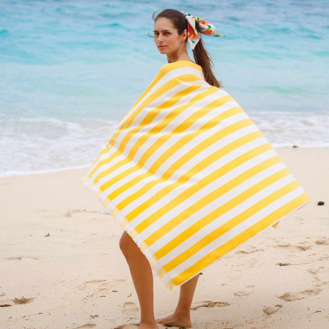 Woman on a beach with a yellow and white striped Green Petition Delmor Ochre branded Beach Towel