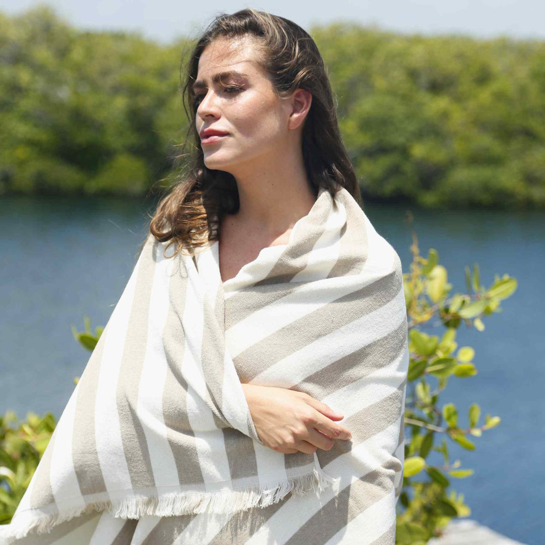 A woman wrapped in a striped 3003 Green Petition Delmor Sand Beige towel standing by a body of water with greenery.