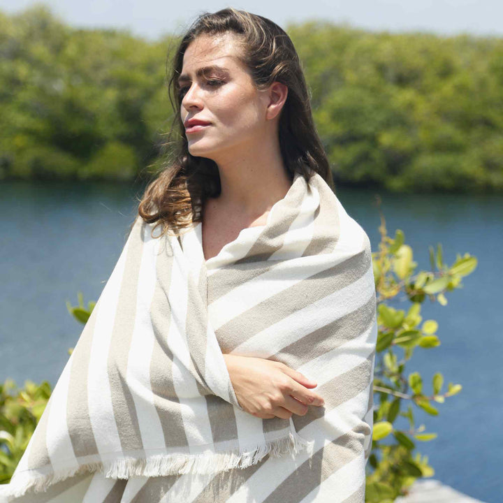 A woman wrapped in a striped 3003 Green Petition Delmor Sand Beige towel standing by a body of water with greenery.