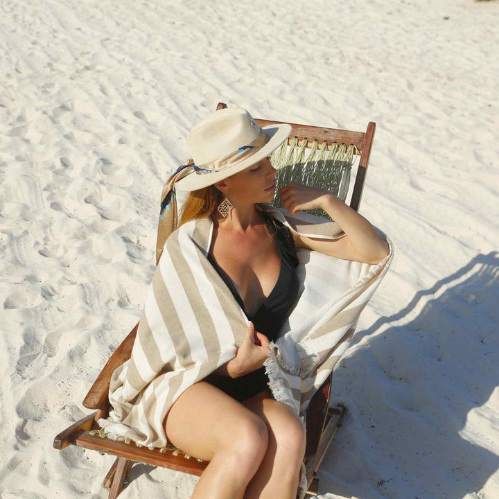 A woman sitting on a beach chair with a Green Petition Delmor Sand Beige towel draped over her, wearing a wide-brimmed hat.