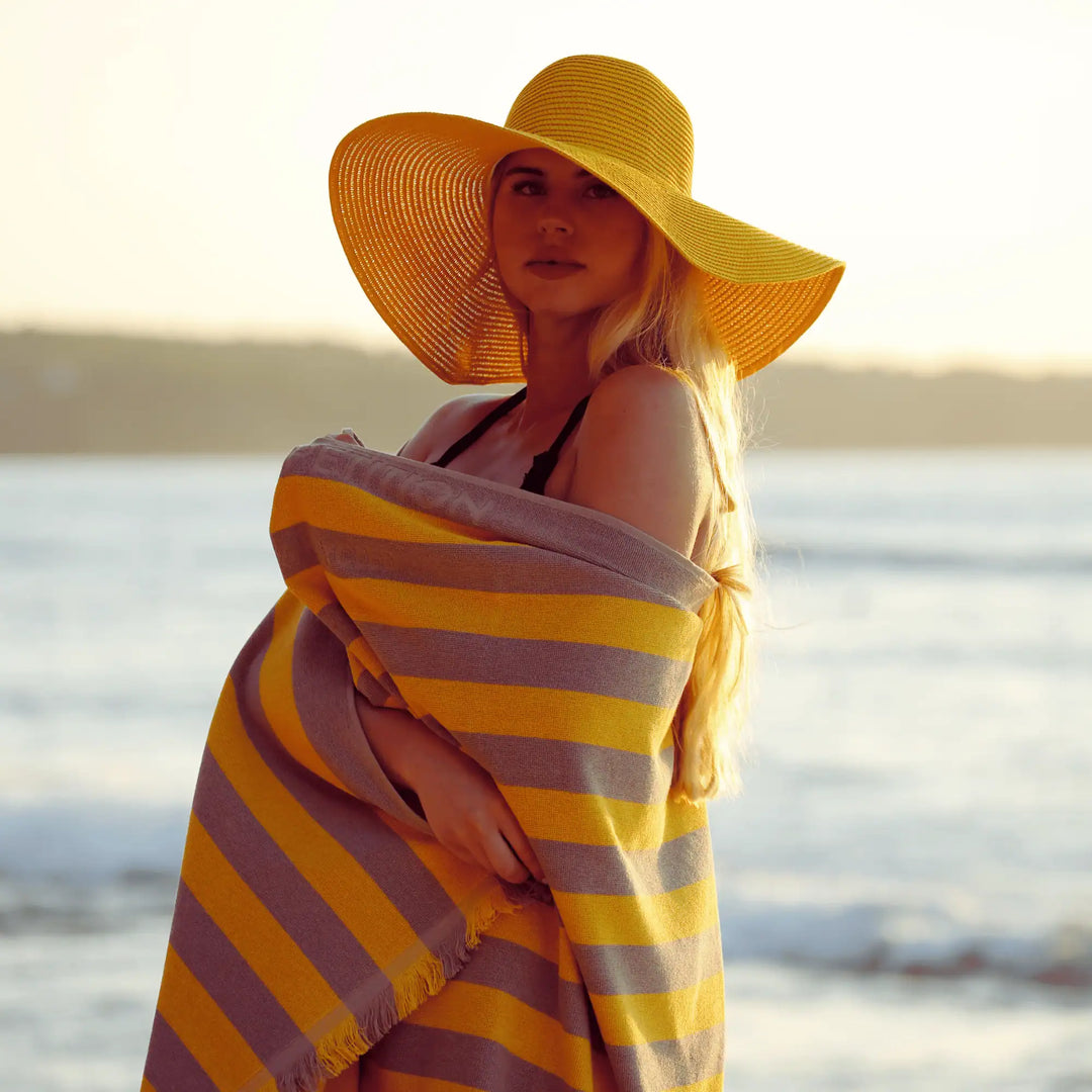 Woman on a beach wearing a yellow hat and "Green Petition Maris Amber" striped towel, with a blurred background of water and sky.