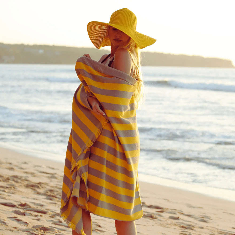Woman on a beach wrapped in a yellow and gray striped "Green Petition Maris Amber" towel, wearing a yellow hat.