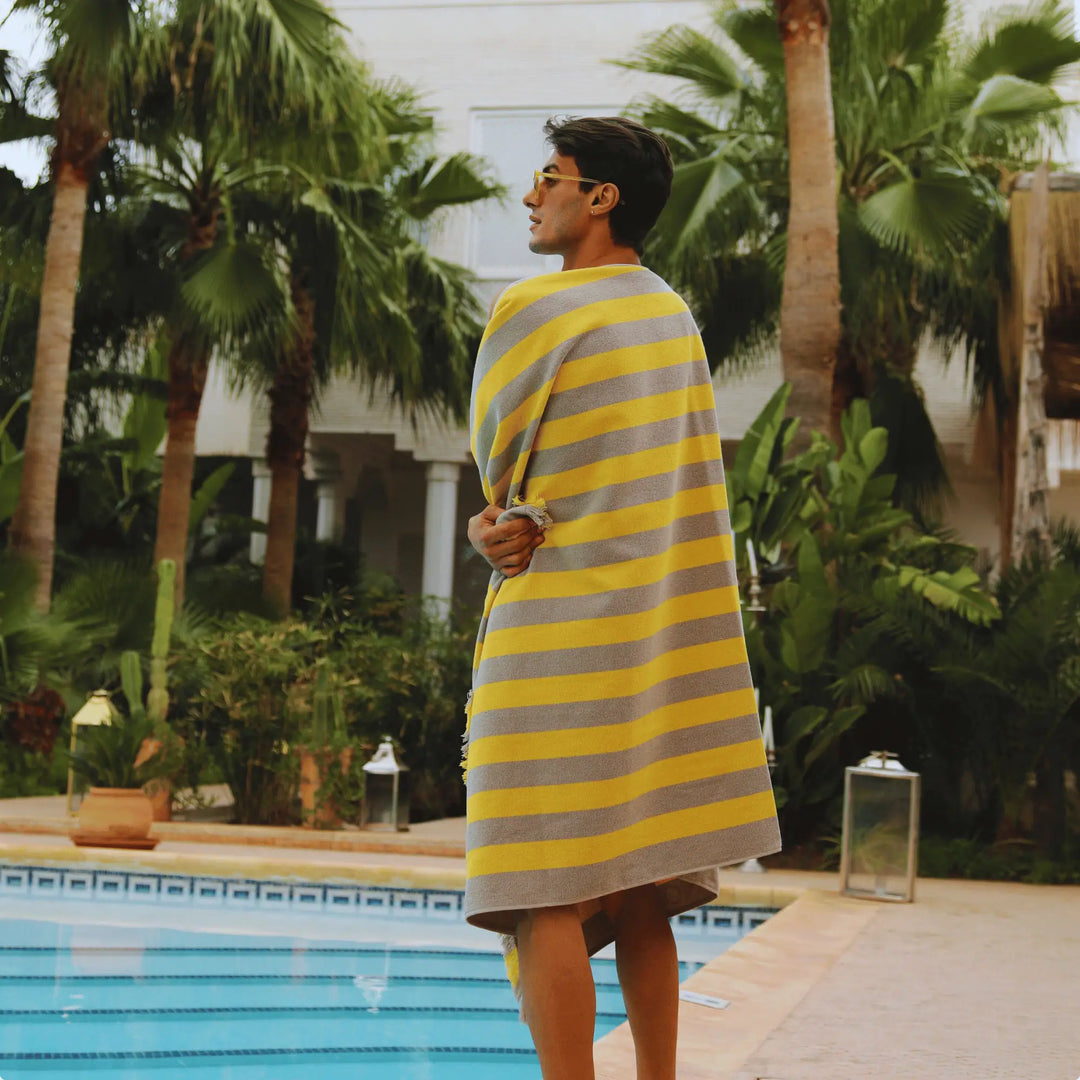 Person wrapped in a yellow and gray striped "Green Petition Maris Amber" towel by a pool with palm trees in the background.