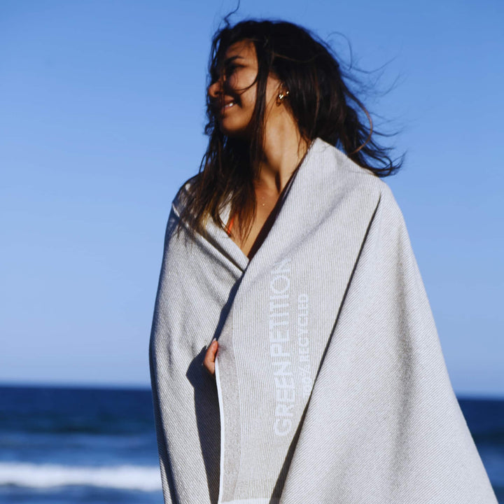 A woman covering her stomach with a grey Green Petition CALM clay towel stands on a beach with an ocean view in the background.