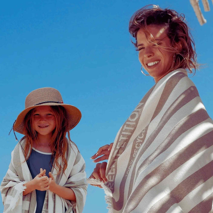 A woman and child wrapped in striped Green Petition Delmor FIT Sand Beige towels against a clear blue sky