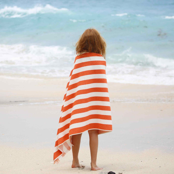 a kid wrapped in an orange and white striped Green Petition Delmor Fit Tangerine Beach Towel towel on a beach