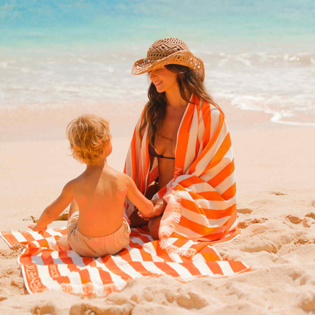 Woman and child sitting on a beach towel at the beach on an orange and white striped Green Petition Delmor Fit Tangerine Beach Towel
