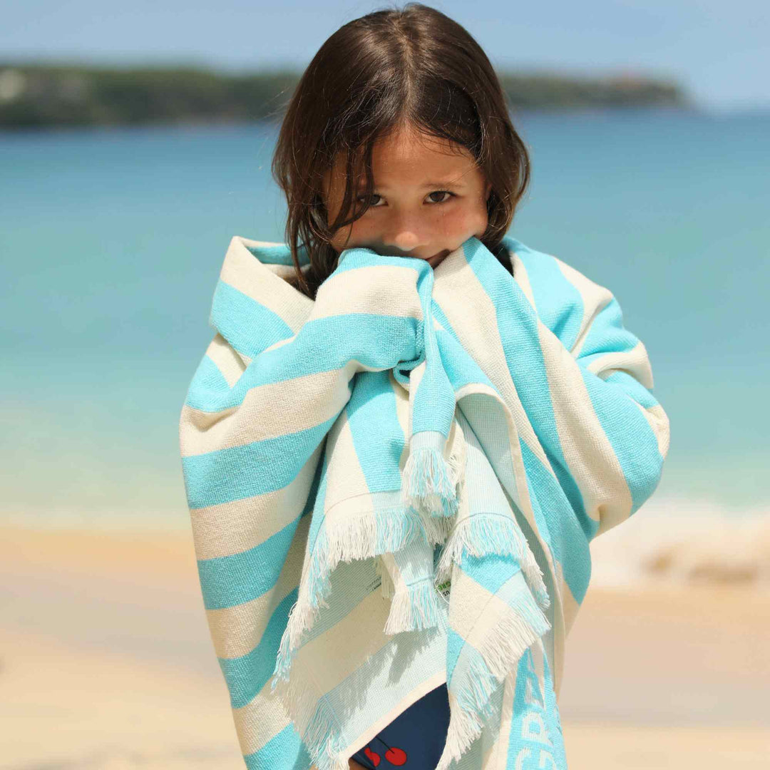 Child wrapped in a blue and white striped Green Petition Delmor Fit Capri towel on a beach