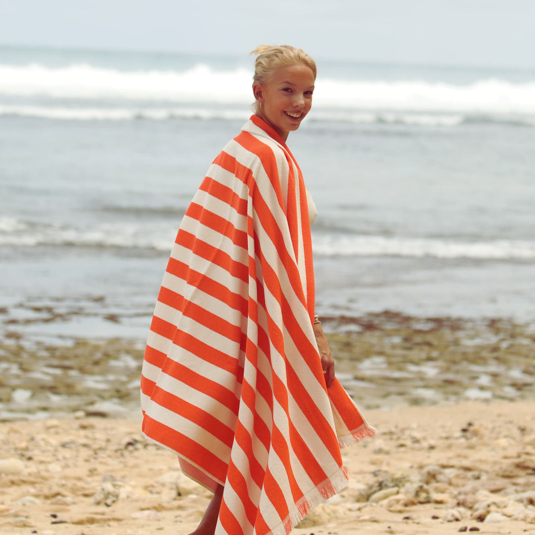 a woman wearing a orange and white striped Green Petition Delmor Tangerine beach towel on a beach
