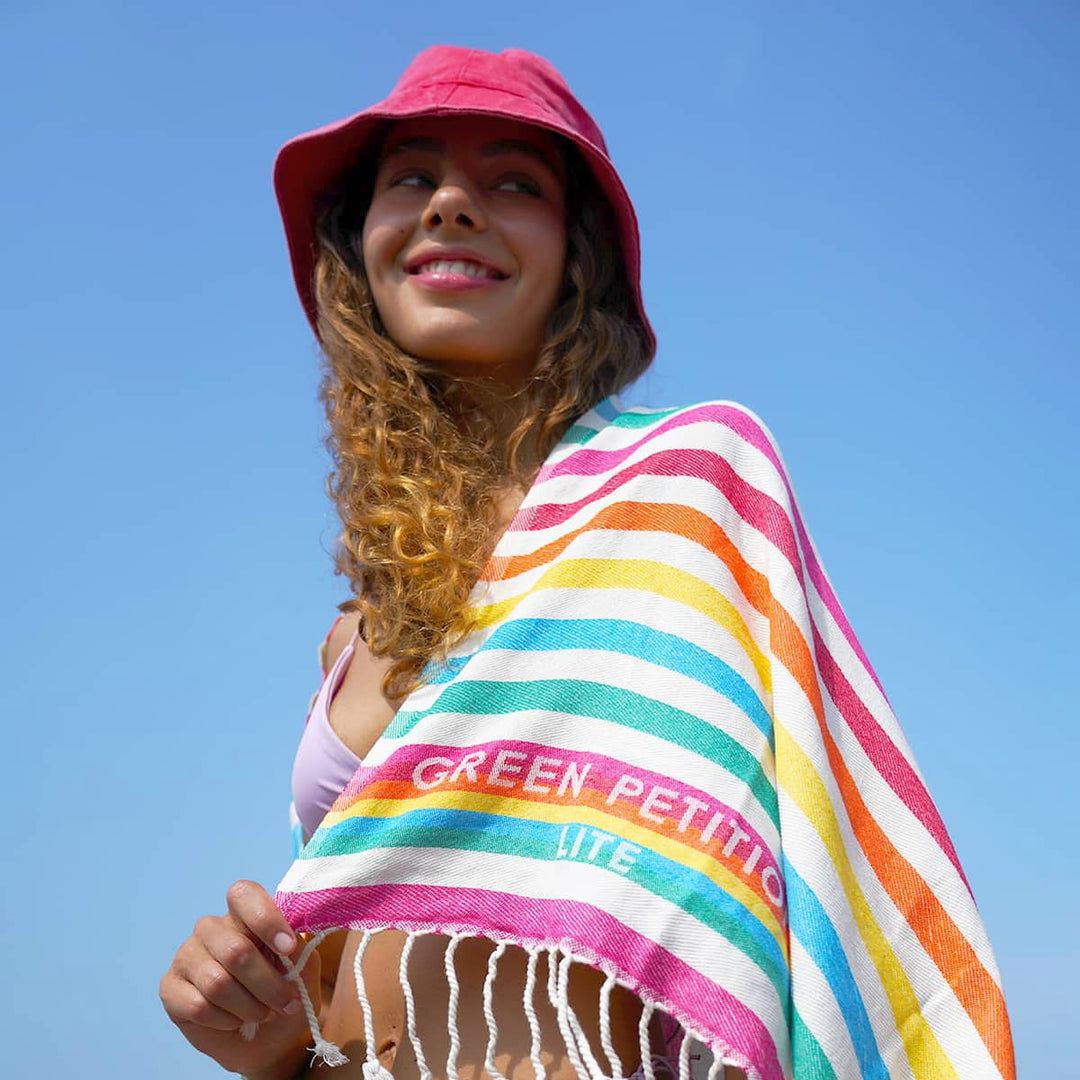 Green Petition recycled dye-free towel in multicolor stripes worn by a woman with a red bucket hat against a clear blue sky, with Green Petition logo woven into the textile.
