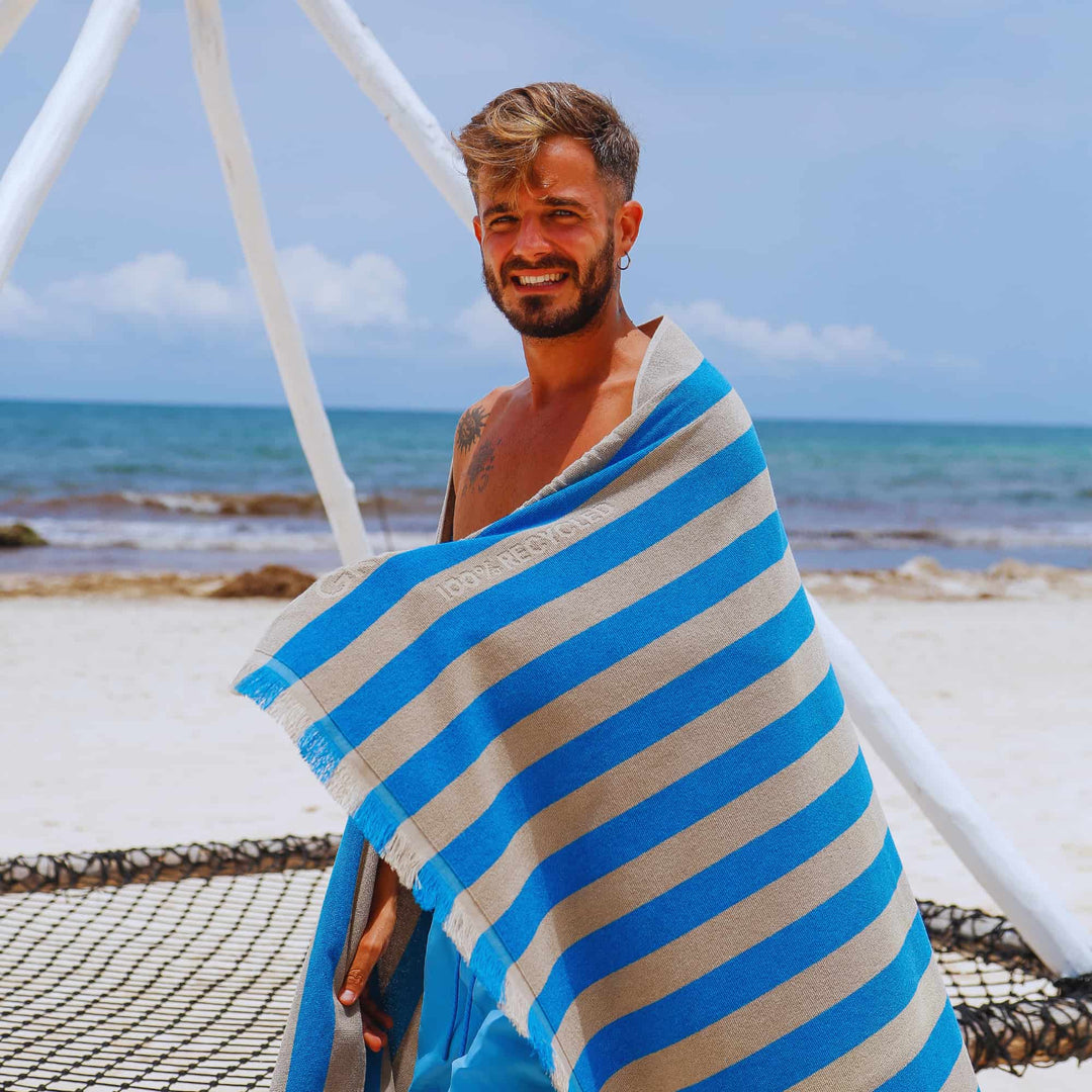 A man wrapped in a blue and beige striped "Green Petition Maris Laguna" Beach Towel on a beach