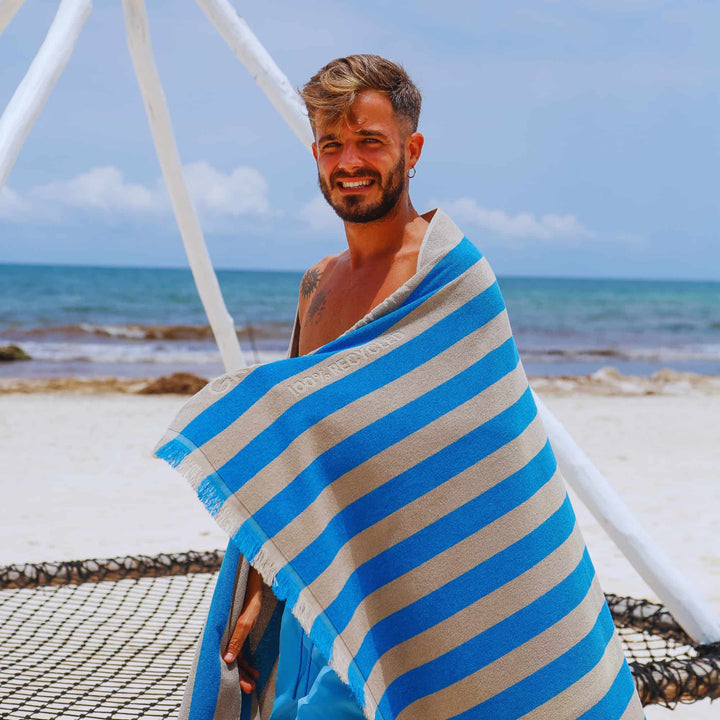 A man wrapped in a blue and beige striped "Green Petition Maris Laguna" Beach Towel on a beach