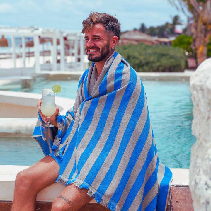 Man wrapped in a blue and white striped "Green Petition Maris Laguna" Beach Towel, holding a drink by a pool.