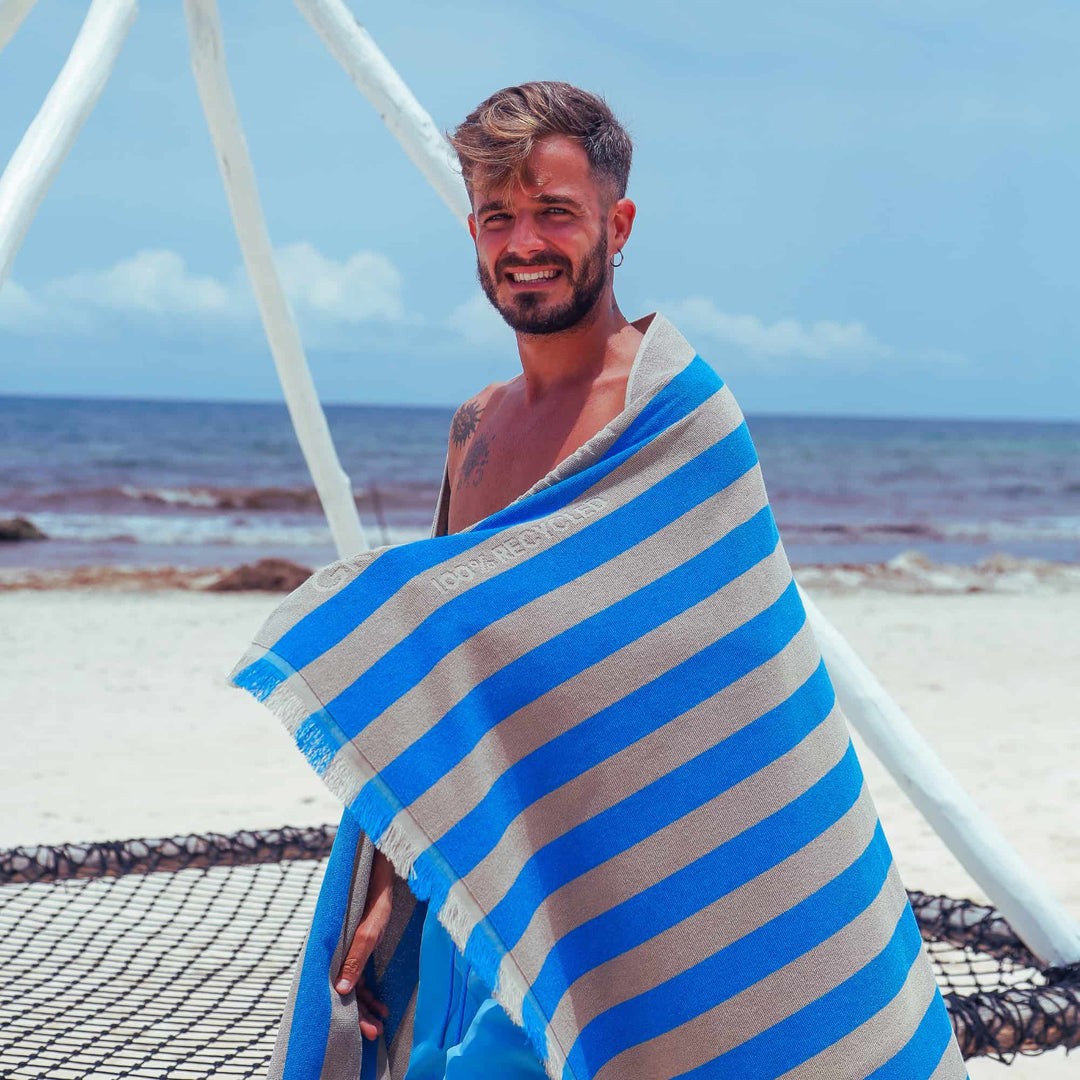 A man wrapped in a blue and gray striped "Green Petition Maris Laguna" Beach Towel  on a beach