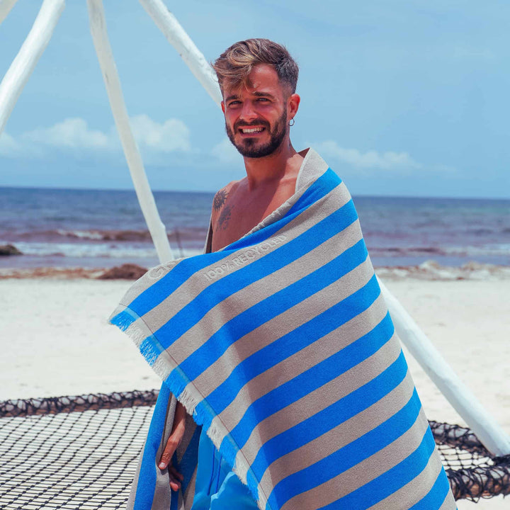 A man wrapped in a blue and gray striped "Green Petition Maris Laguna" Beach Towel  on a beach