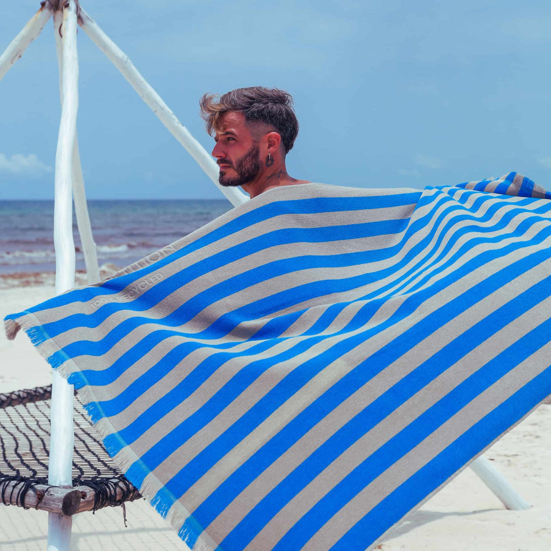 Man sitting on a beach chair with a blue and white striped "Green Petition Maris Laguna" Beach Towel  draped over him, ocean in the background
