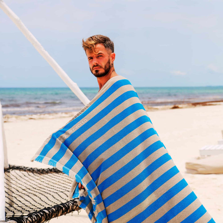 Man on a beach wrapped in a blue and white striped "Green Petition Maris Laguna" Beach Towel 
