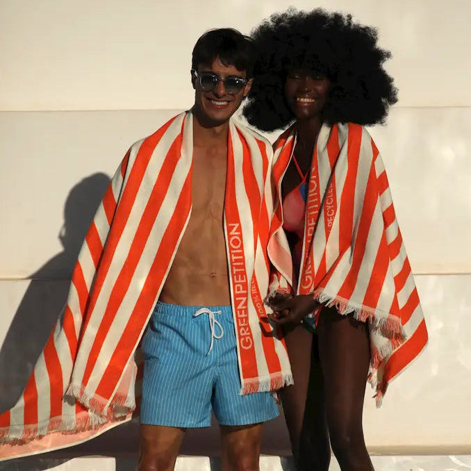 Two people standing in water, they  wrapped in a tangerine and cream striped  Green Petition 100% Recycled towel, against a neutral background.