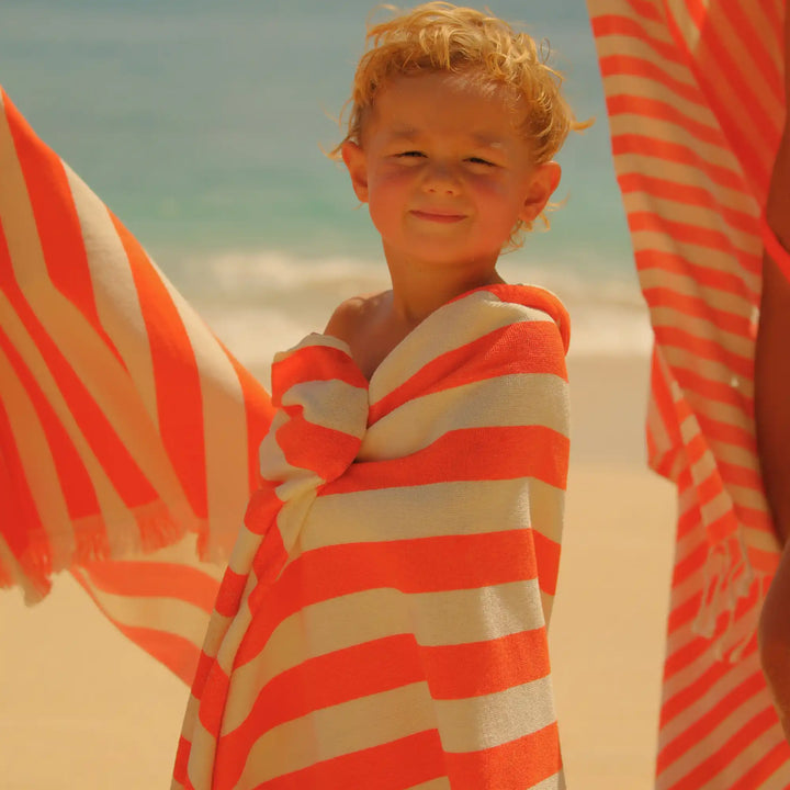 A young child wrapped in a bright orange and white striped "Green Petition Tangerine beach towel" on a sandy beach with the ocean in the background.