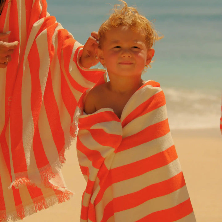 A child wrapped in a bright orange and white striped "Green Petition" brand "Tangerine" color beach towel on a sandy beach, with ocean water in the background.