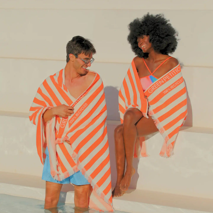 Two models wrapped in bright orange and white striped Green Planet beach towels, standing and sitting in shallow pool water.