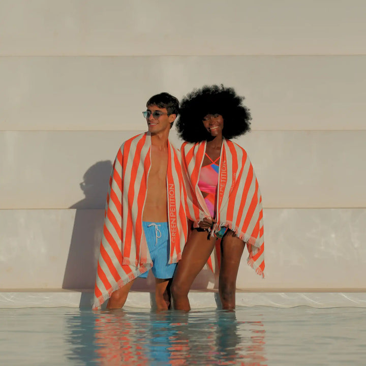A man and a woman standing in shallow water in front of a white wall, both wrapped in orange and white striped beach towels with the brand name &