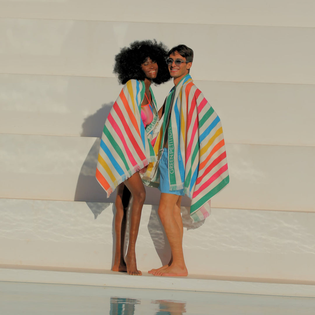 Two models standing on the edge of a pool, wrapped in colorful striped beach towels with the brand name &