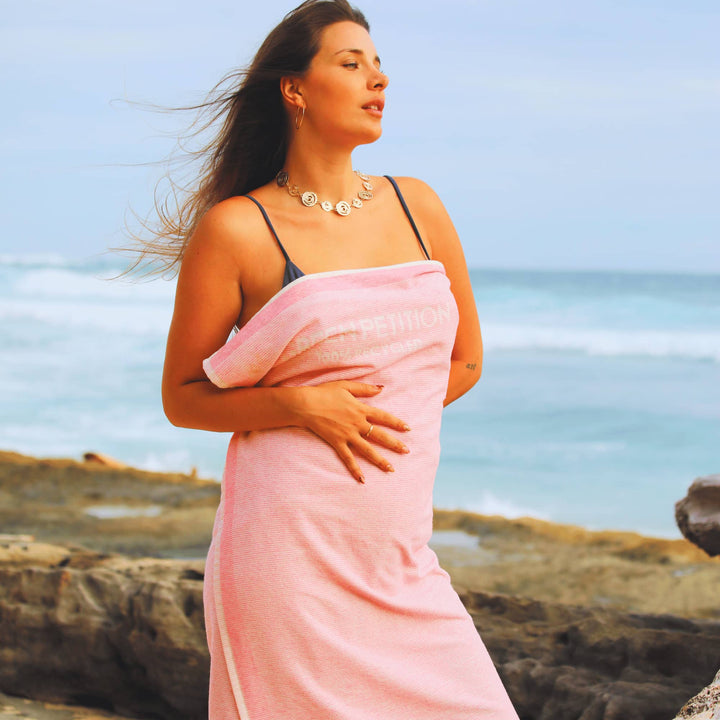 Woman wrapped in a Pink striped Green Petition Calm Candy beach towel on a rocky beach with ocean in the background
